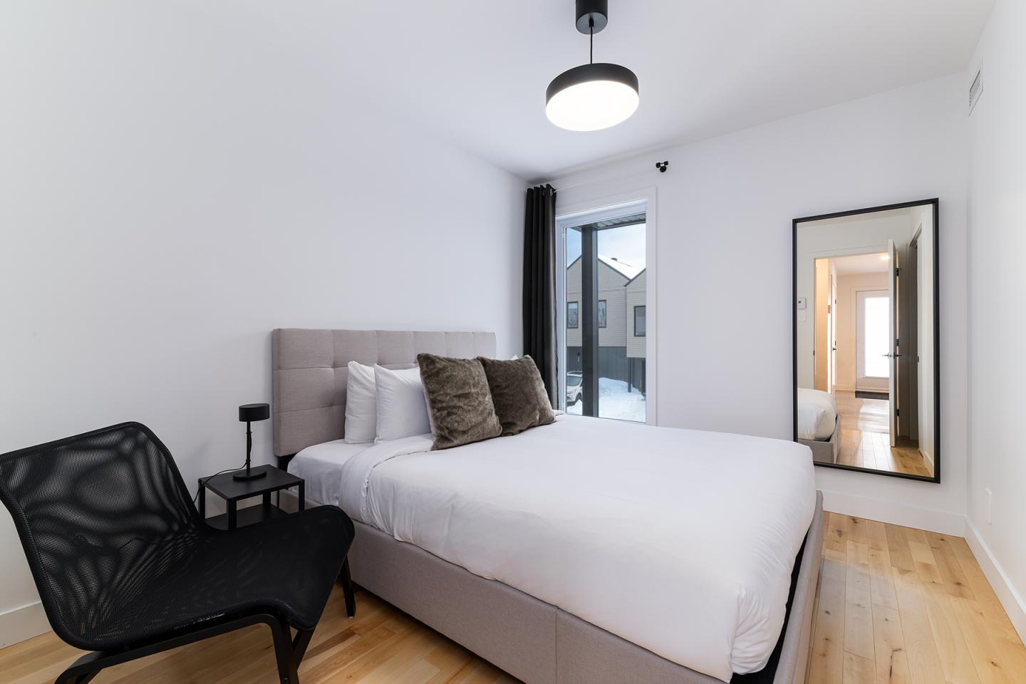 Chambre moderne avec lit double, chaise noire et miroir sur mur blanc.