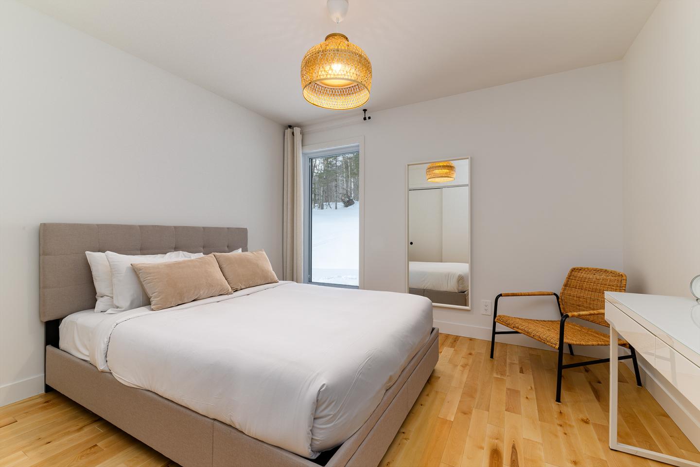 Chambre moderne avec lit double, chaise en osier, bureau blanc et fenêtre sur paysage enneigé.