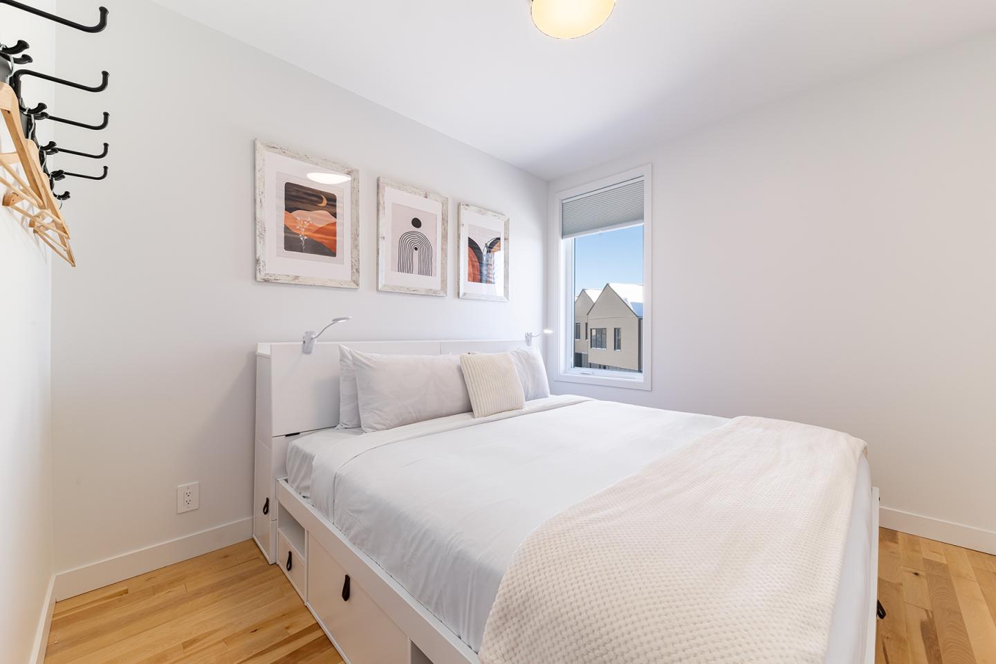 Petite chambre blanche avec un lit double, cadre minimaliste et fenêtre ensoleillée.