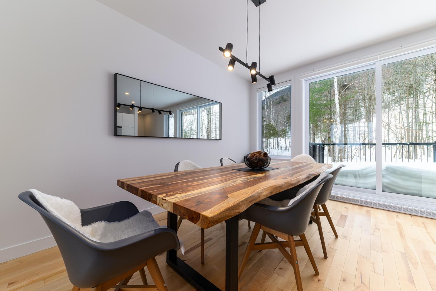 Salle à manger moderne avec table en bois, chaises grises, grande fenêtre, lumière naturelle.