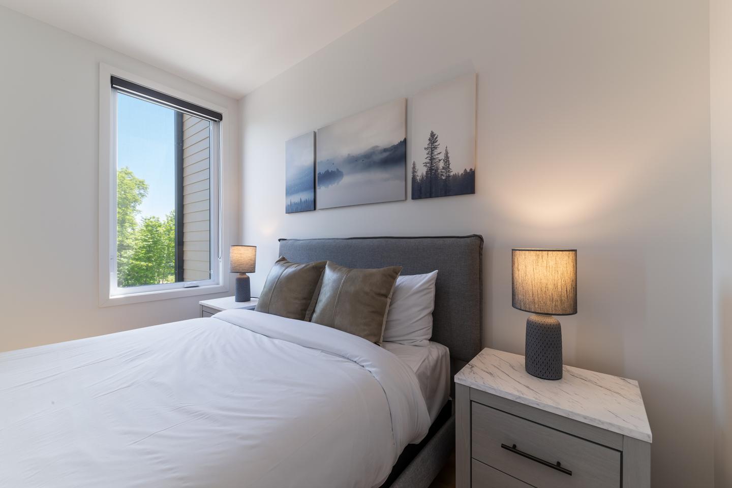 Chambre lumineuse, lit gris, lampes allumées, fenêtre avec vue sur la verdure.