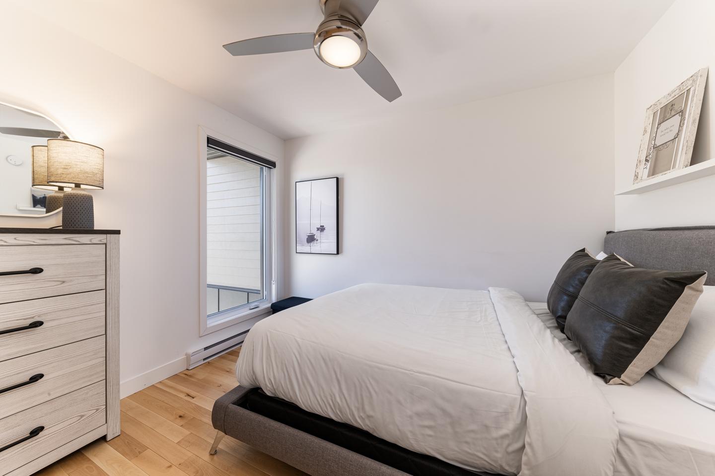 Chambre moderne avec lit double, ventilateur, commode et grande fenêtre.