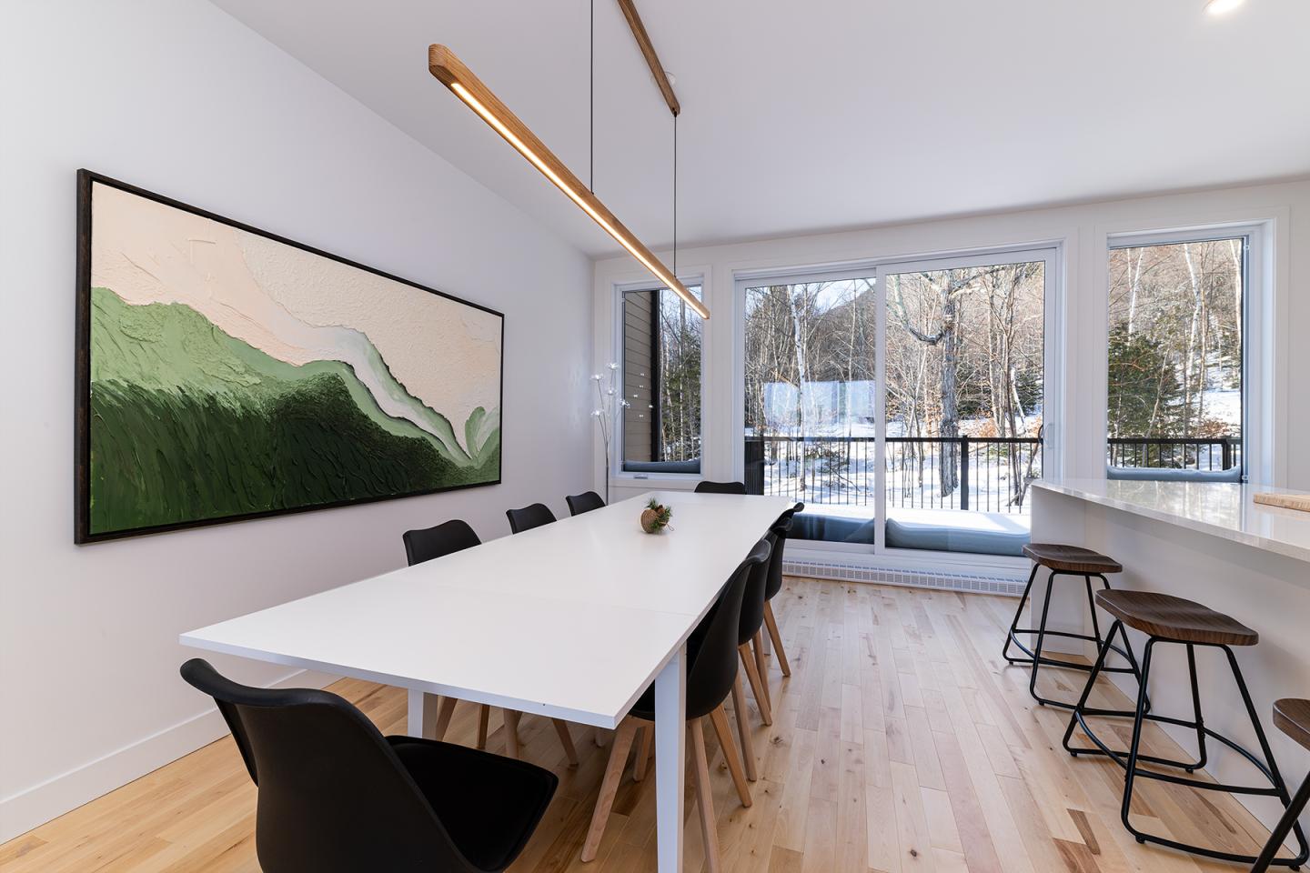 Salle à manger moderne, grande table blanche, chaises noires, tableau abstrait, baie vitrée.