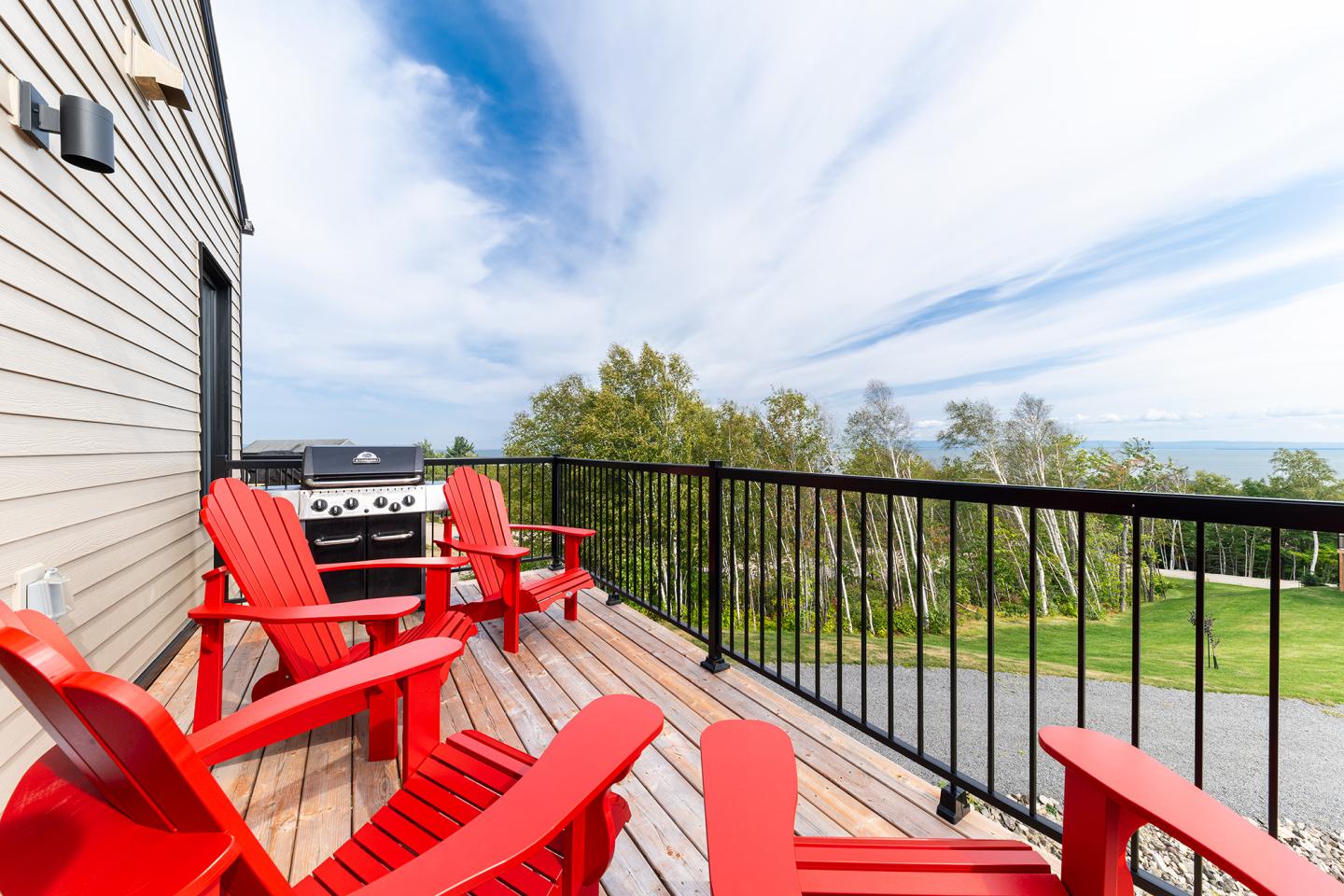 Terrasse en bois avec chaises rouges et barbecue, vue sur jardin verdoyant.