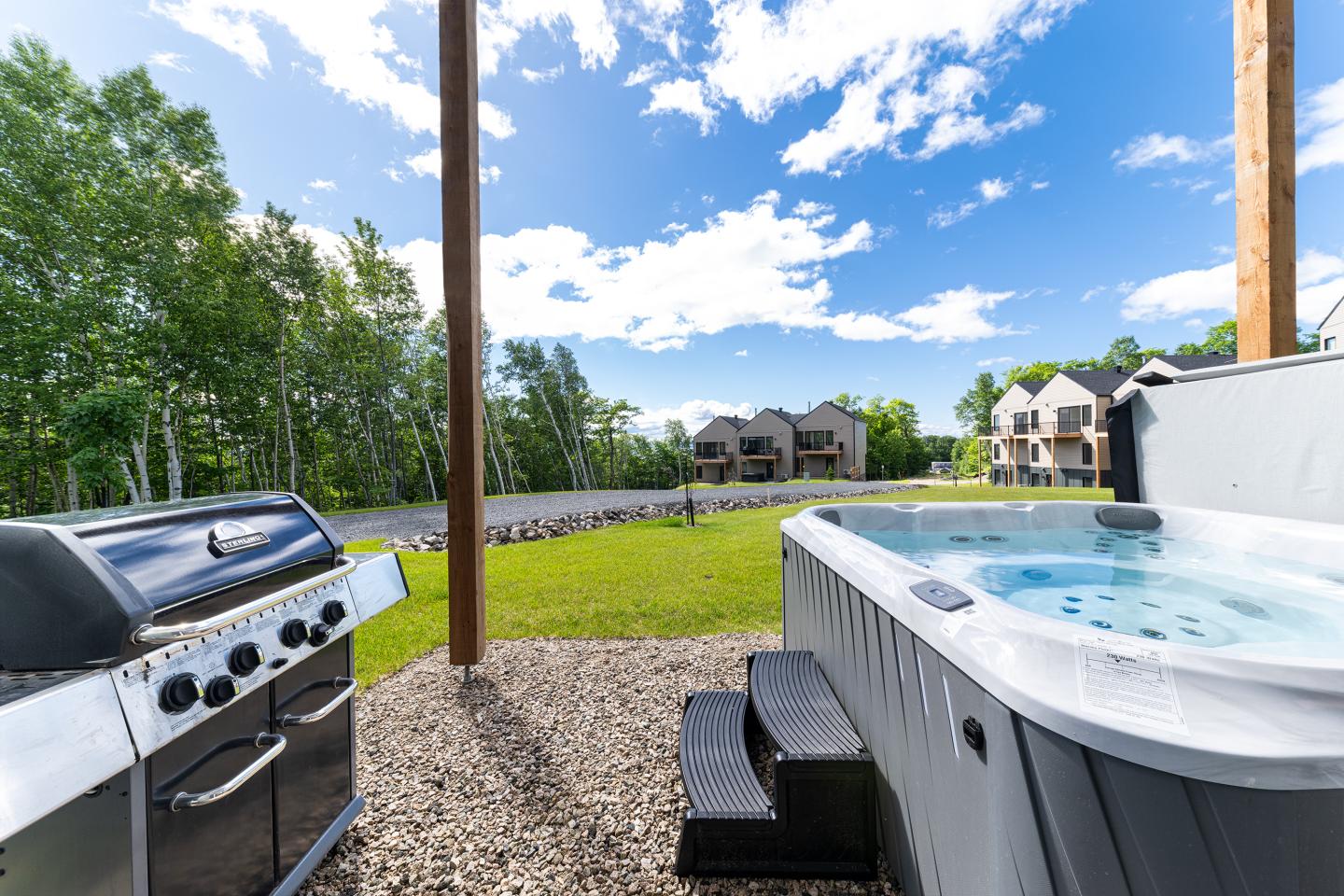 Jacuzzi et barbecue sur terrasse avec vue sur jardin et maisons, ciel bleu.
