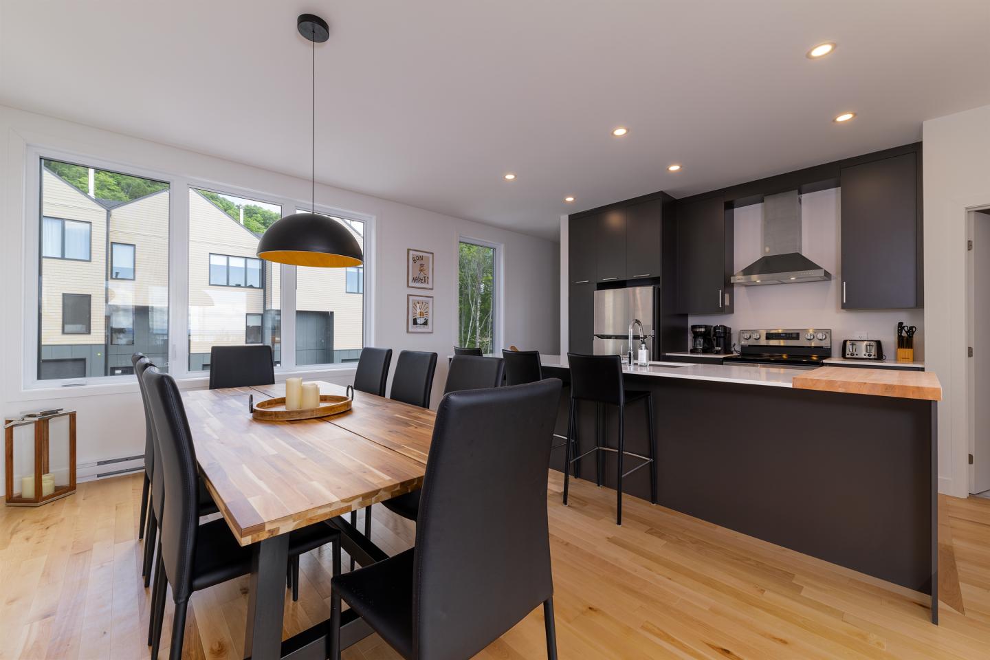 Salle à manger moderne avec table en bois, chaises noires, cuisine ouverte à l'arrière.