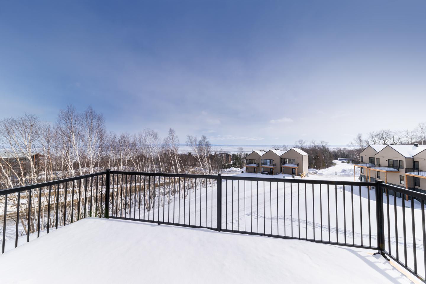 Balcon enneigé avec vue sur des arbres et maisons lointaines sous ciel bleu.