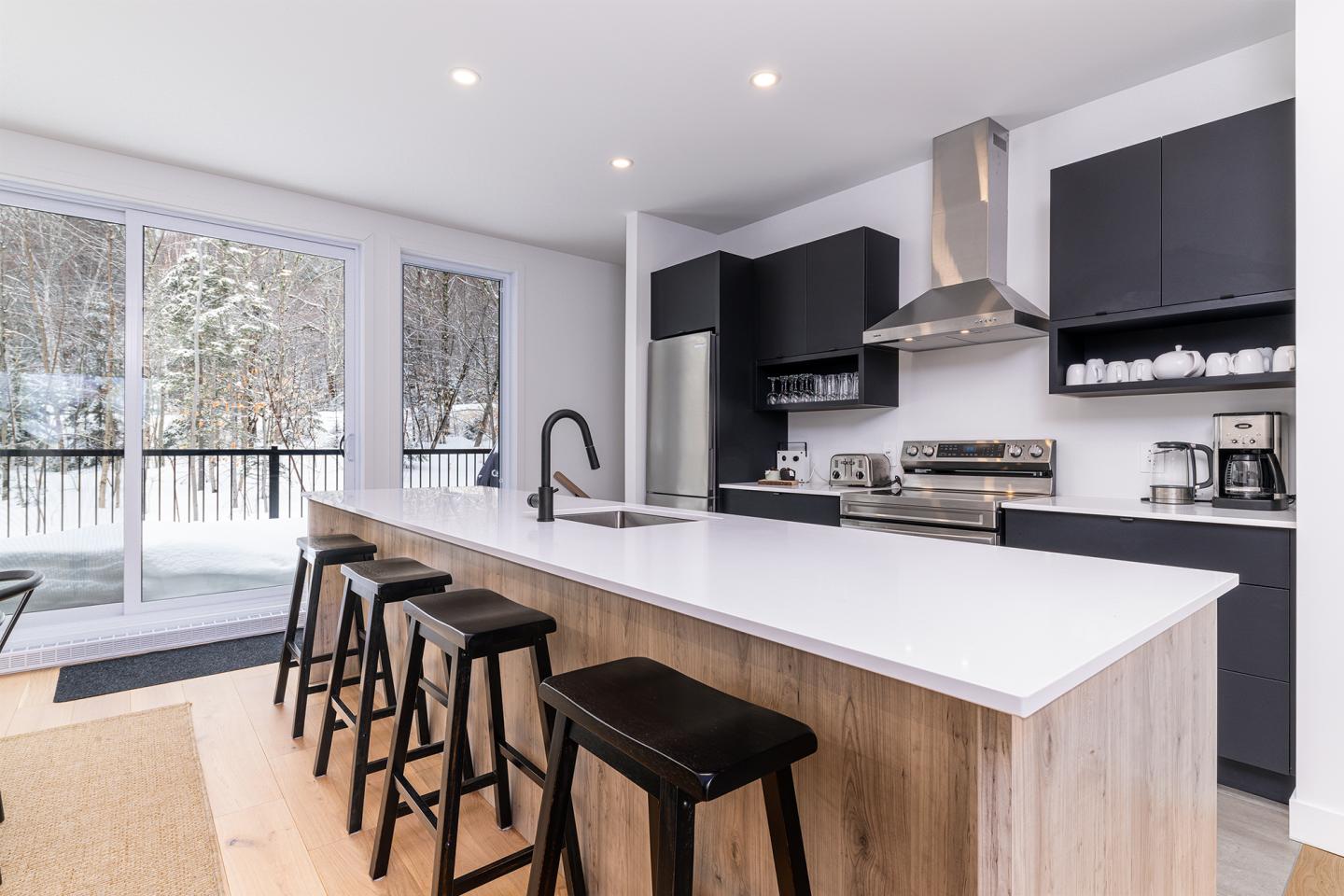 Cuisine moderne avec îlot central, chaises noires, grandes fenêtres, électroménagers en inox.