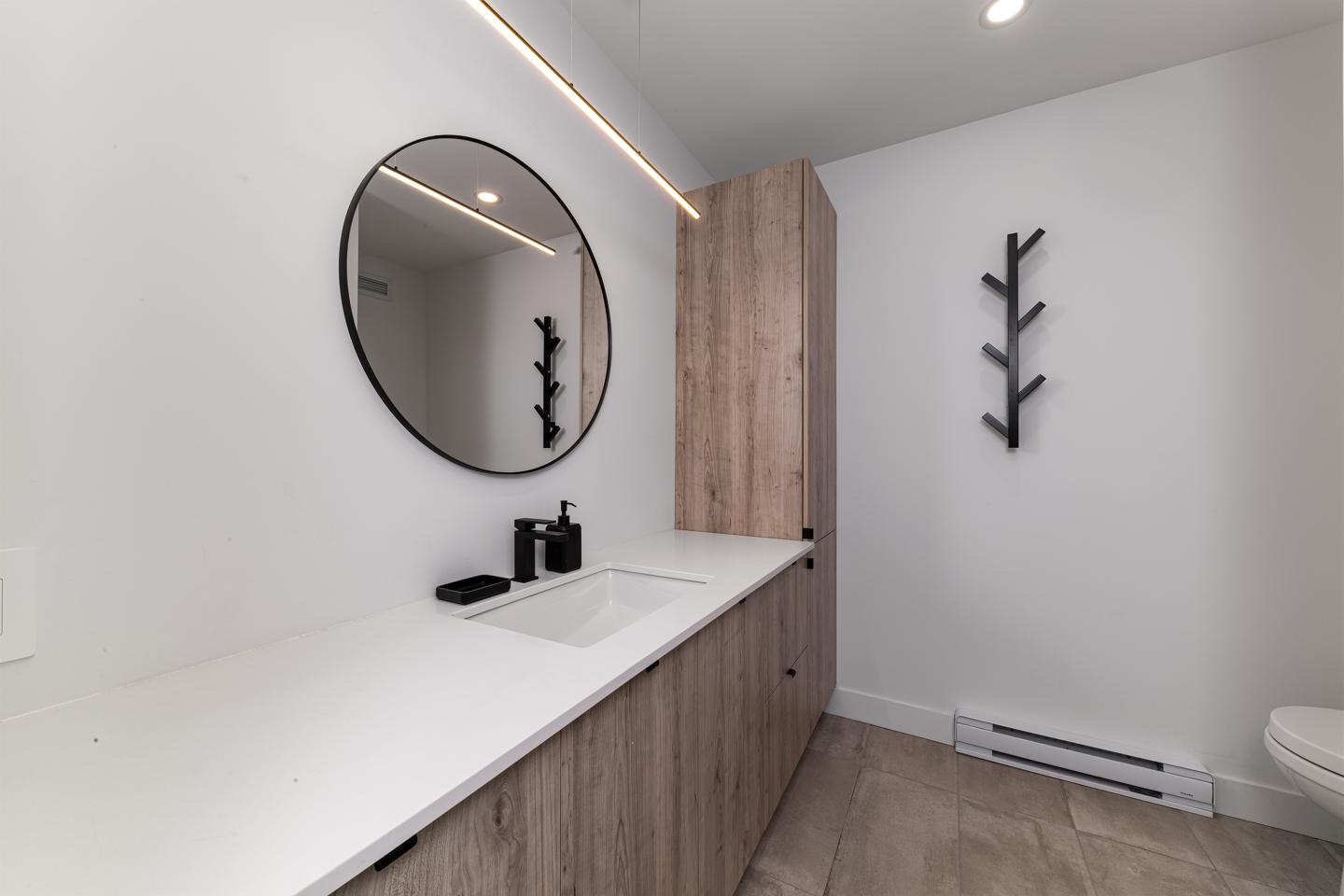 Salle de bain moderne avec miroir rond, comptoir blanc et meuble en bois.