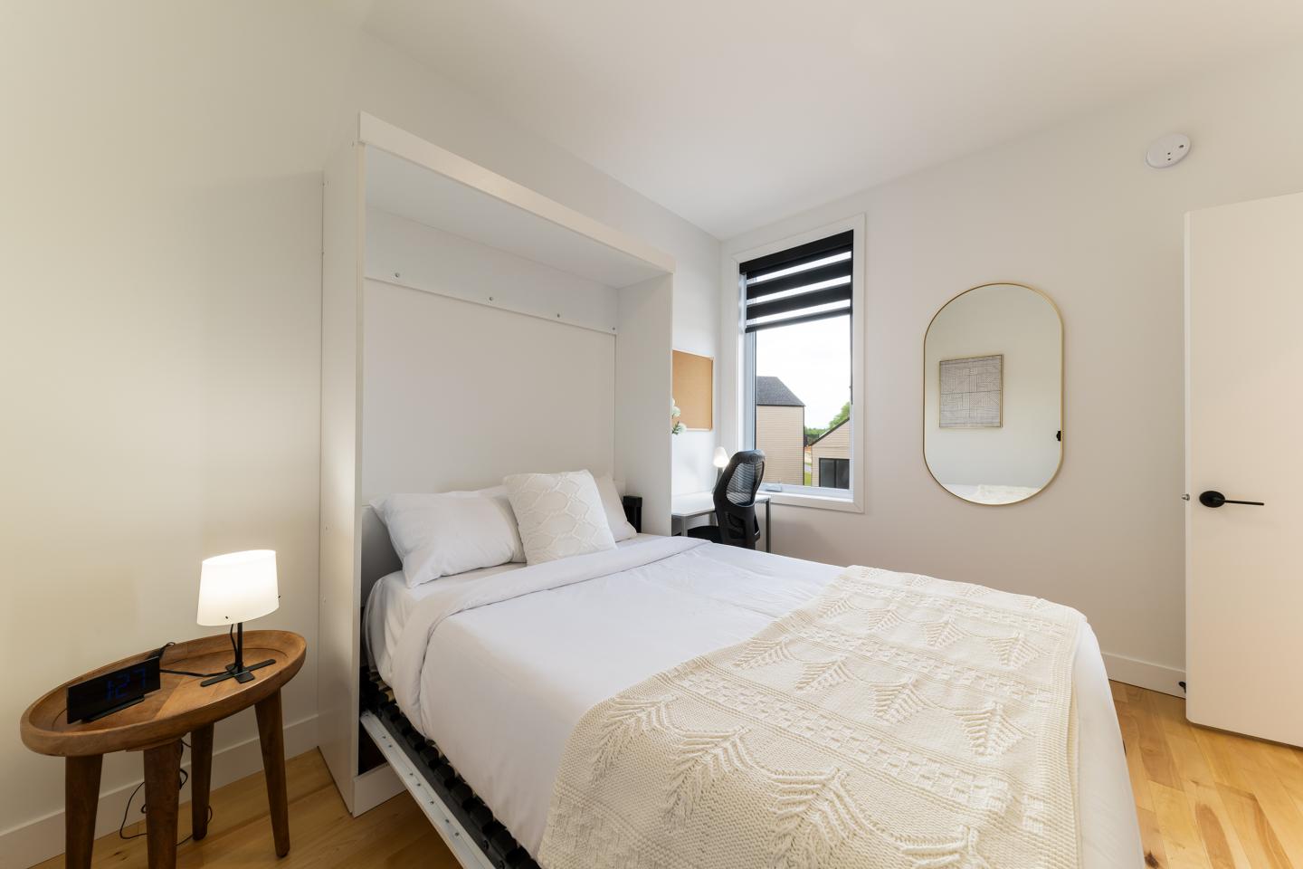 Chambre moderne avec lit blanc, miroir ovale, et lampe sur une table en bois.
