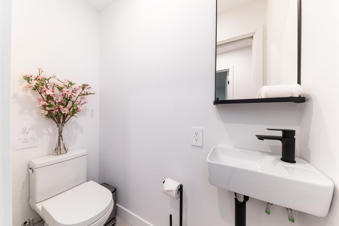 Petite salle de bain blanche avec lavabo, miroir et fleurs roses sur le réservoir des WC.