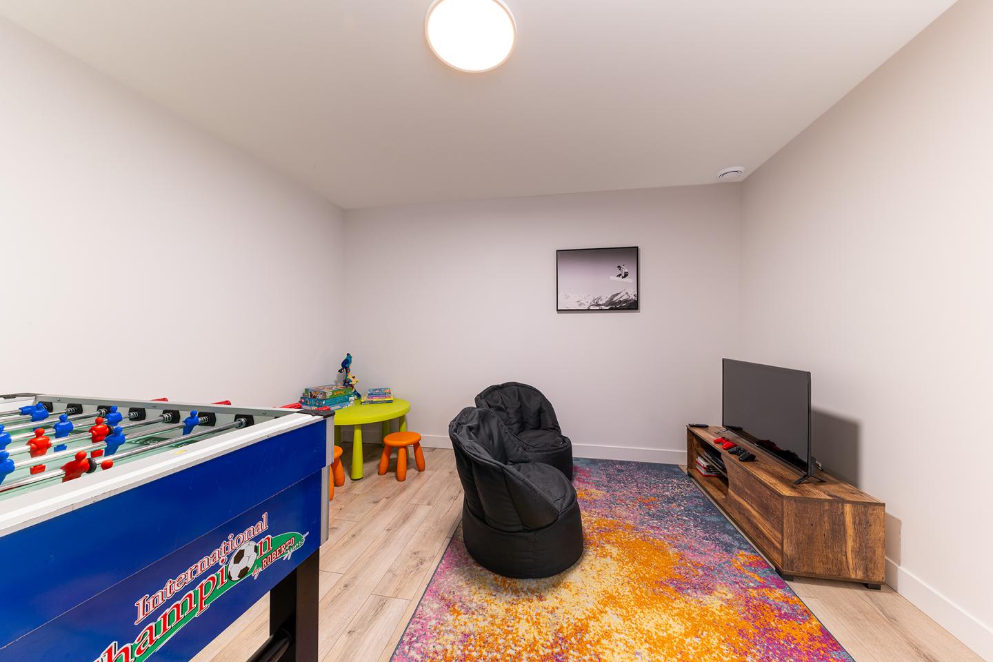 Salle de jeux avec baby-foot, télé, fauteuil et tapis coloré.