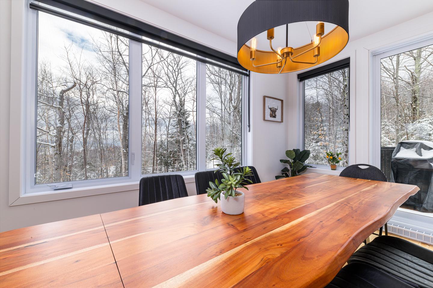 Salle à manger lumineuse avec table en bois et grandes fenêtres donnant sur des arbres enneigés.