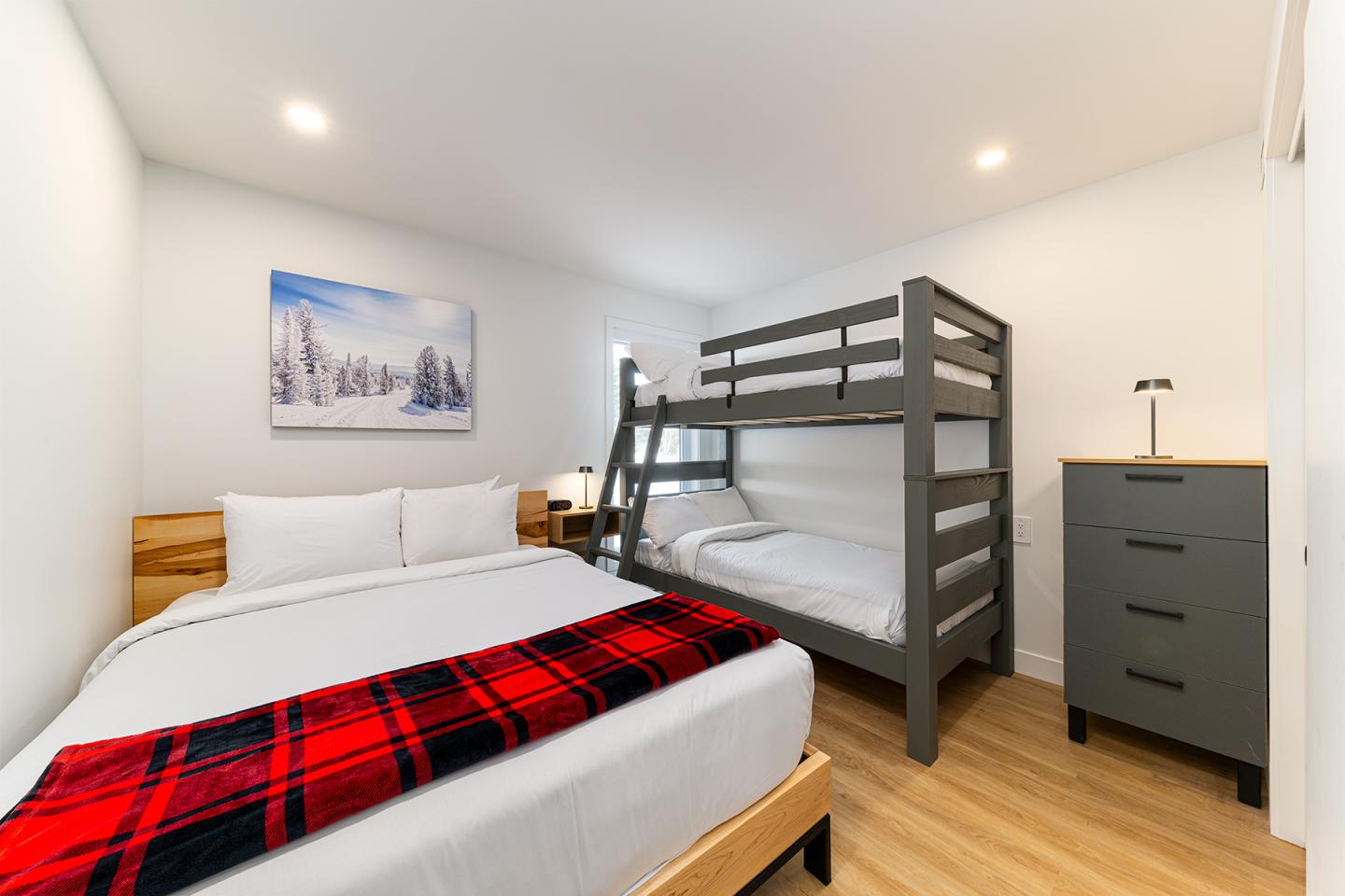 Chambre lumineuse avec lit double, lit superposé gris et commode noire.