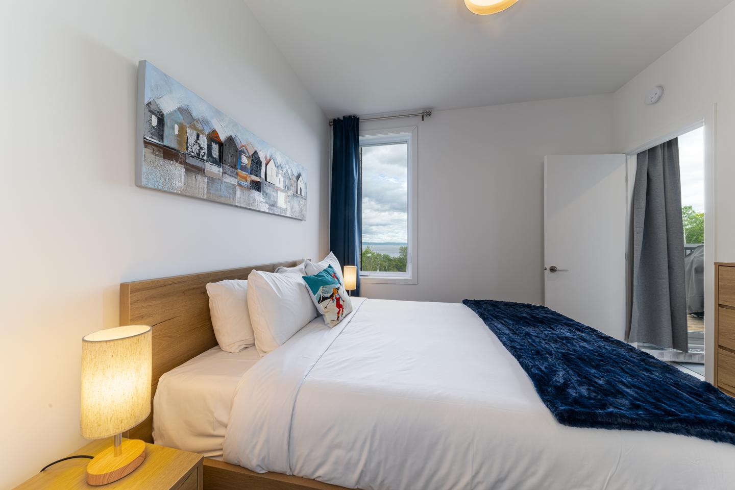Chambre moderne avec lit double, couverture bleue, lampe et fenêtre ouverte.