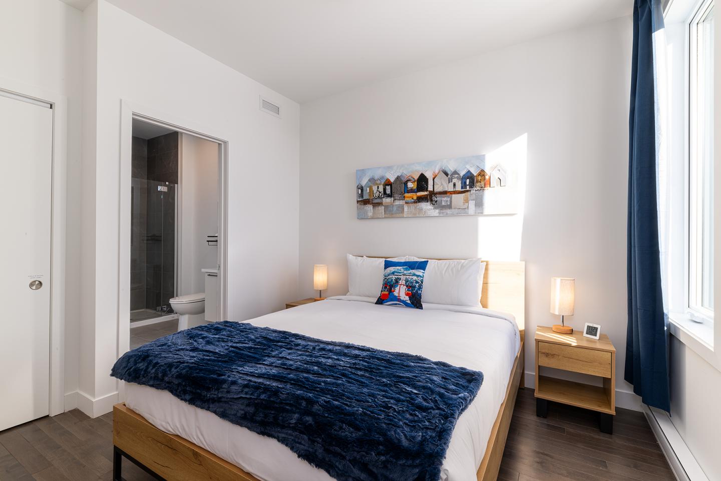 Chambre lumineuse avec grand lit, couverture bleu foncé, tableau mural, et salle de bain attenante.