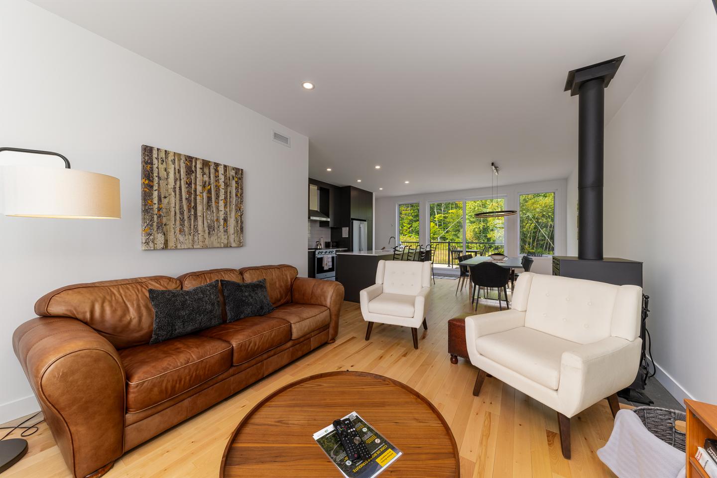 Salon moderne avec canapé en cuir, fauteuils blancs, vue sur cuisine et grandes fenêtres.