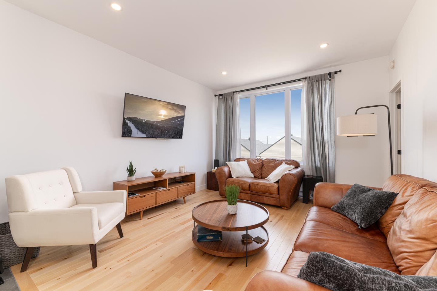 Salon moderne avec canapé en cuir, fauteuil blanc, et TV murale.