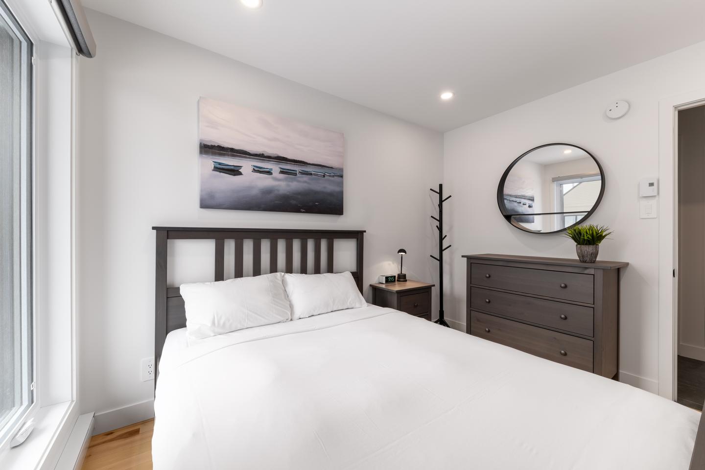 Chambre lumineuse avec lit blanc, commode, et miroir rond.