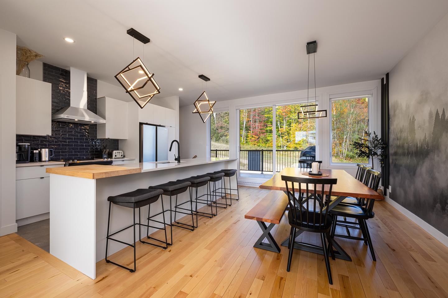 Cuisine moderne avec îlot, tabourets, table en bois, et grandes fenêtres donnant sur jardin.