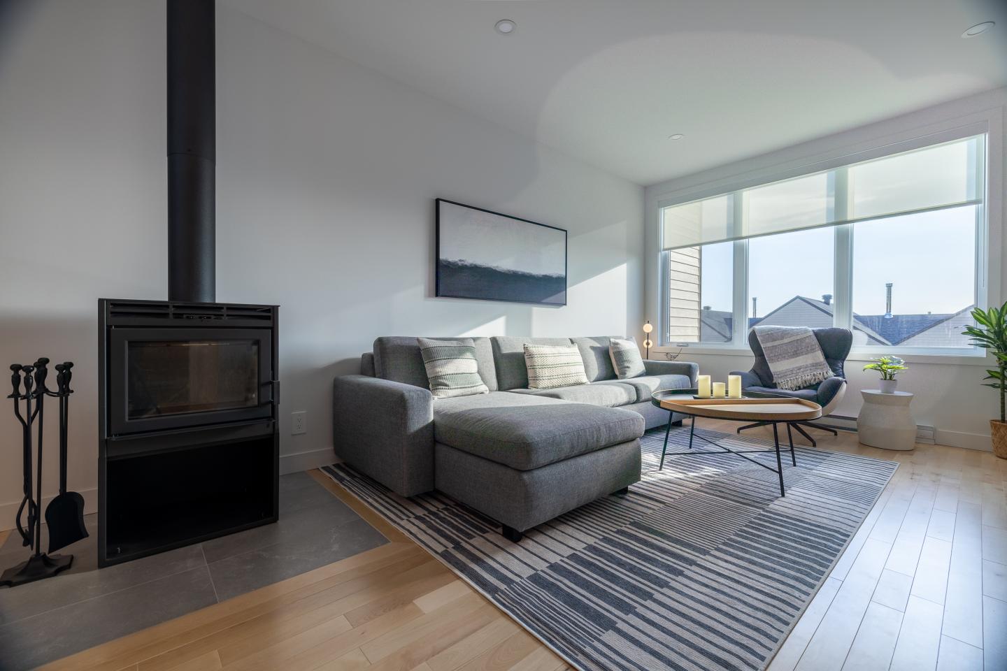 Salon moderne avec canapé gris, poêle à bois et grande fenêtre lumineuse.