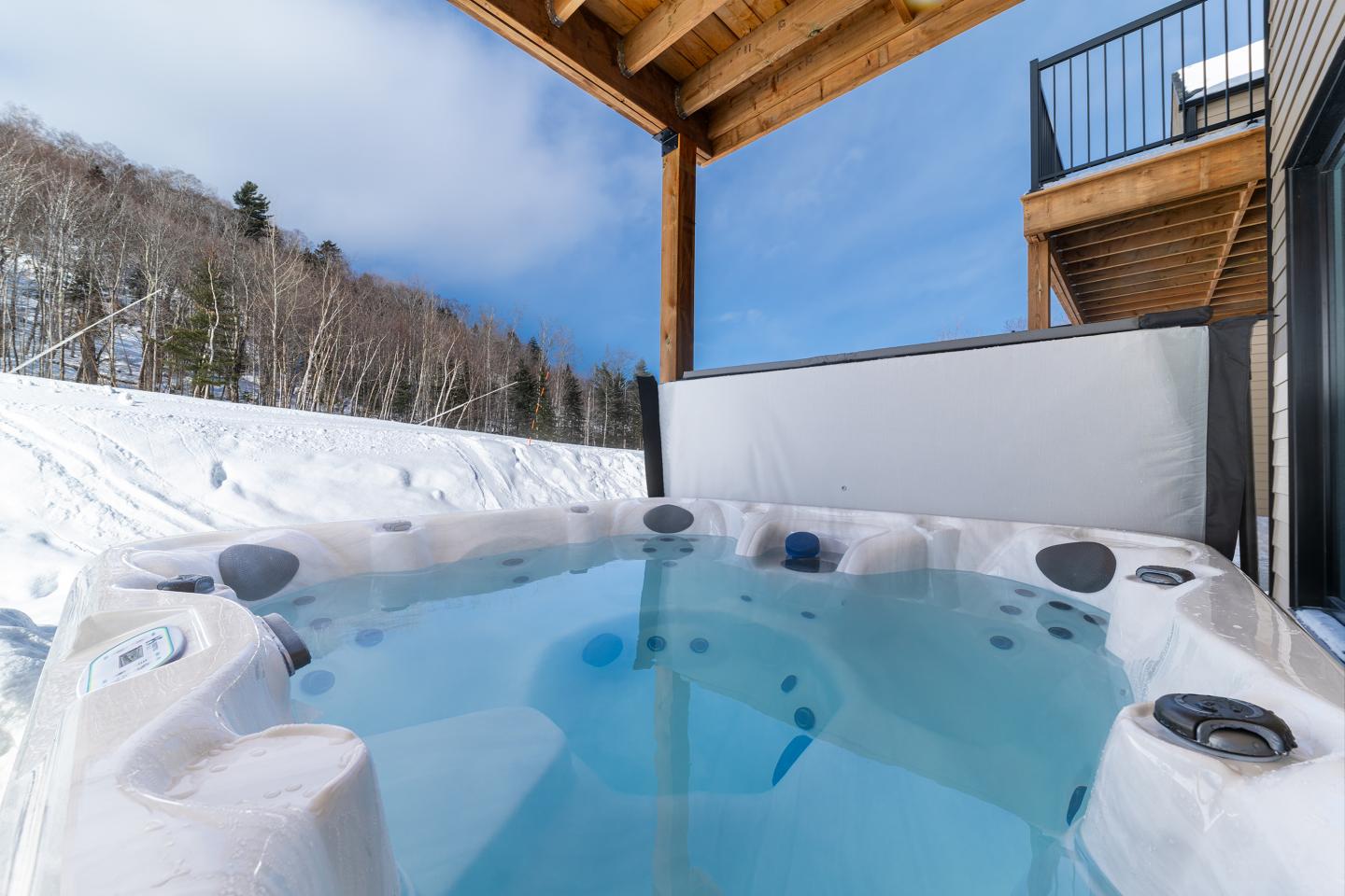 Jacuzzi extérieur sous terrasse, neige et forêt en arrière-plan, ciel bleu.