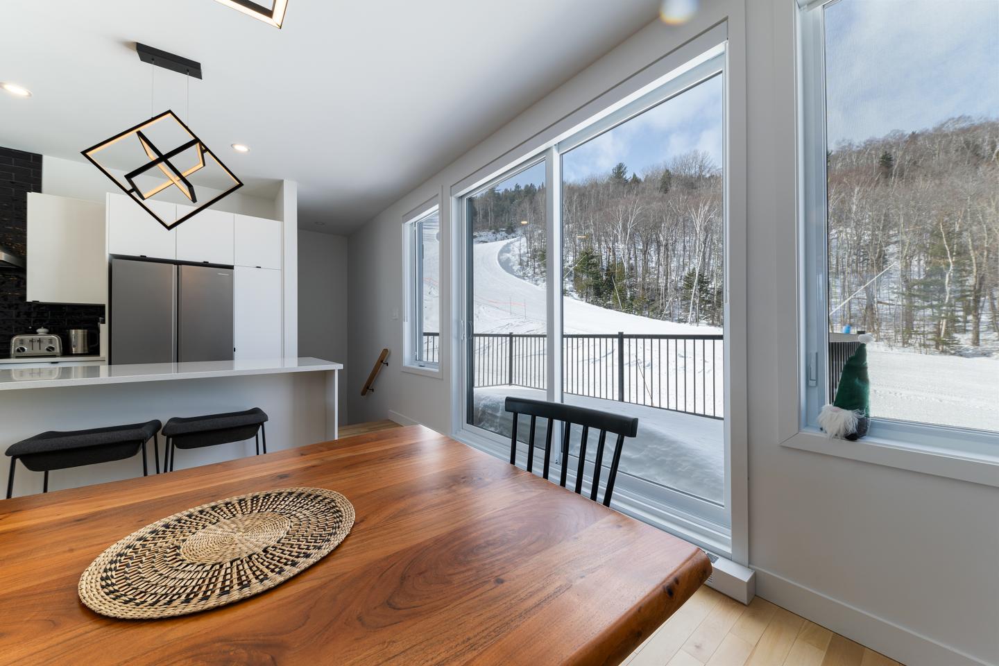 Intérieur lumineux avec grande fenêtre vue sur neige. Table bois et cuisine moderne.
