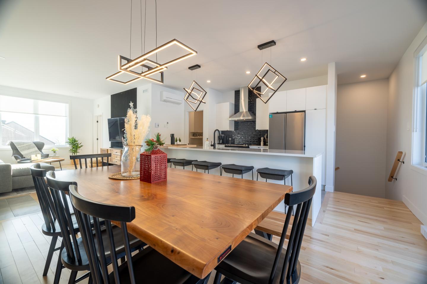 Cuisine moderne et lumineuse avec table en bois, chaises noires et éclairage géométrique.