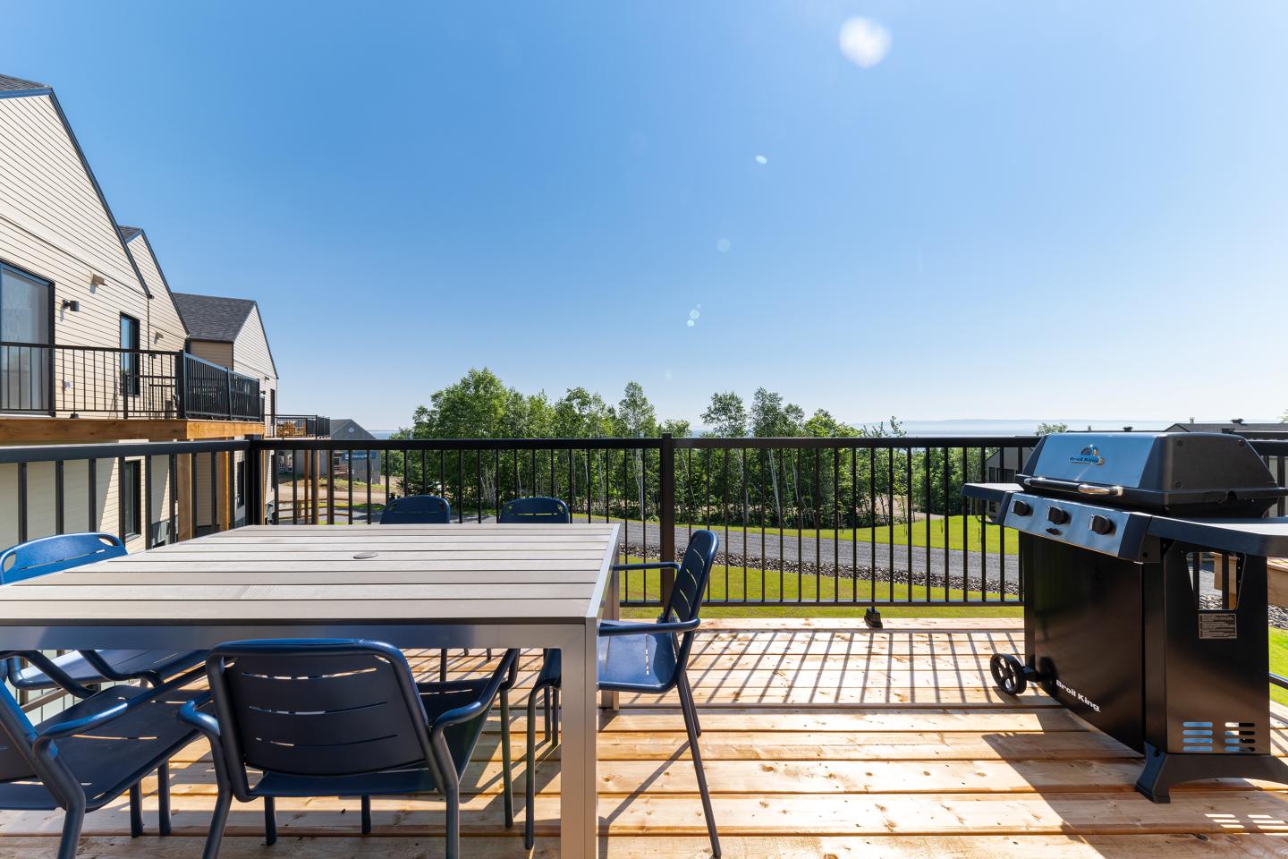 Terrasse ensoleillée avec table, chaises bleues et barbecue, vue sur jardin verdoyant.