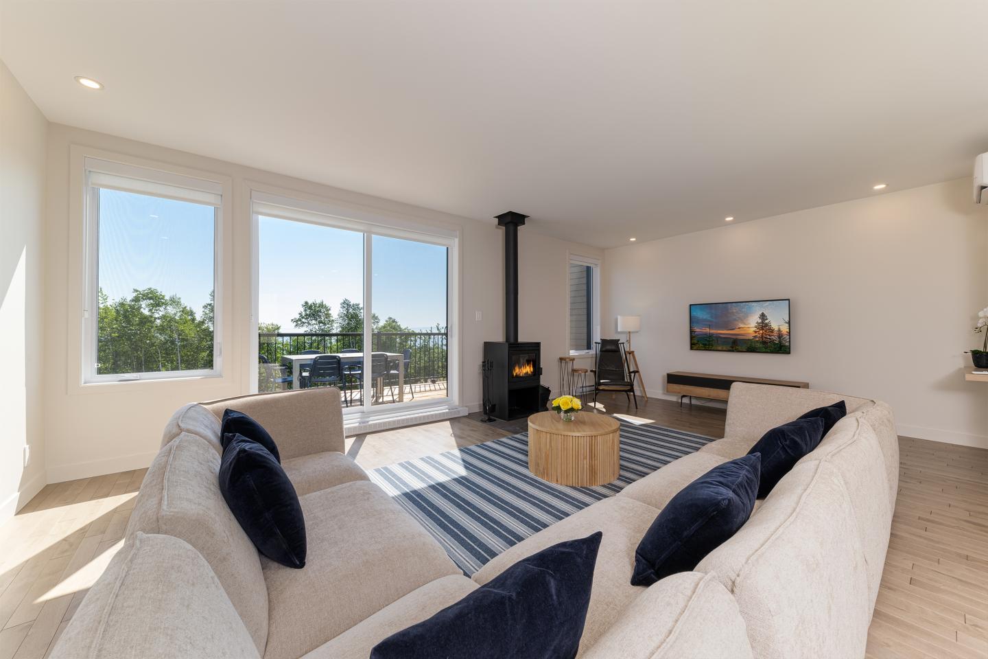 Salon lumineux avec canapé beige, poêle à bois, tapis rayé et grande fenêtre.