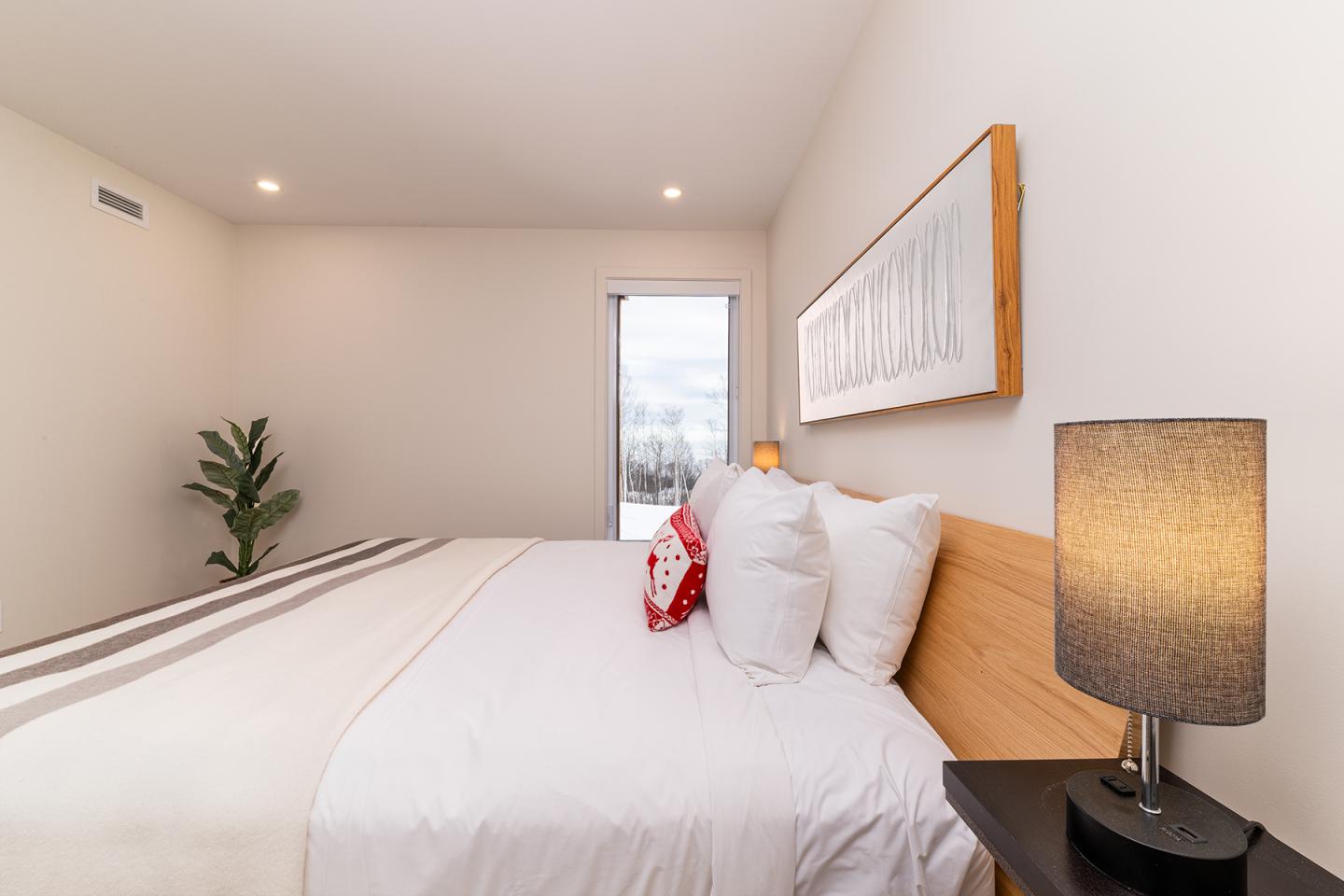 Chambre moderne avec grand lit, lampe allumée, plante verte et fenêtre.
