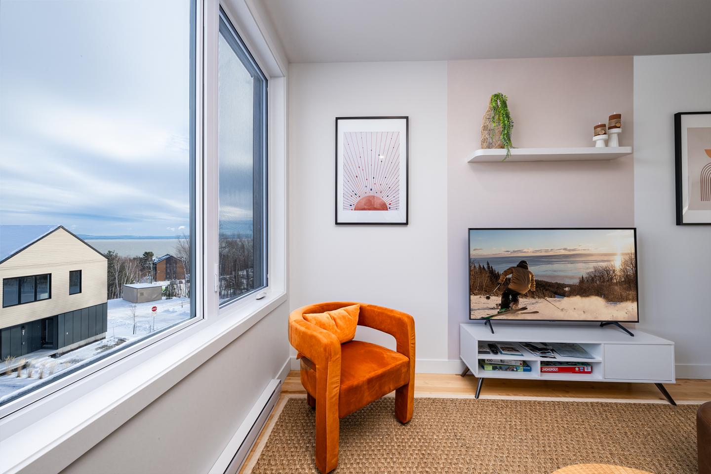 Salon moderne avec fauteuil orange, grande fenêtre et télé montrant paysage enneigé.