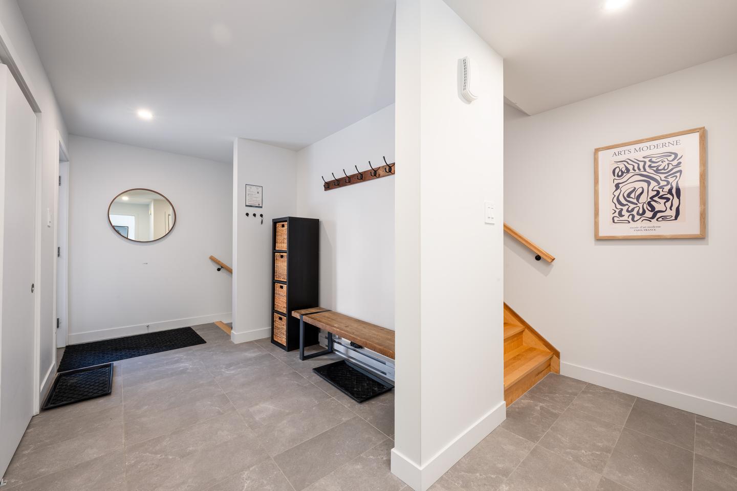 Entrée moderne avec banc en bois, miroir rond et escalier, murs blancs.