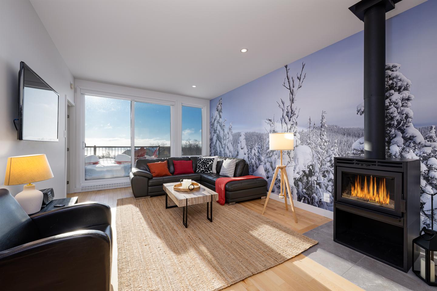 Salon moderne avec cheminée, canapé noir, et vue sur paysage enneigé.