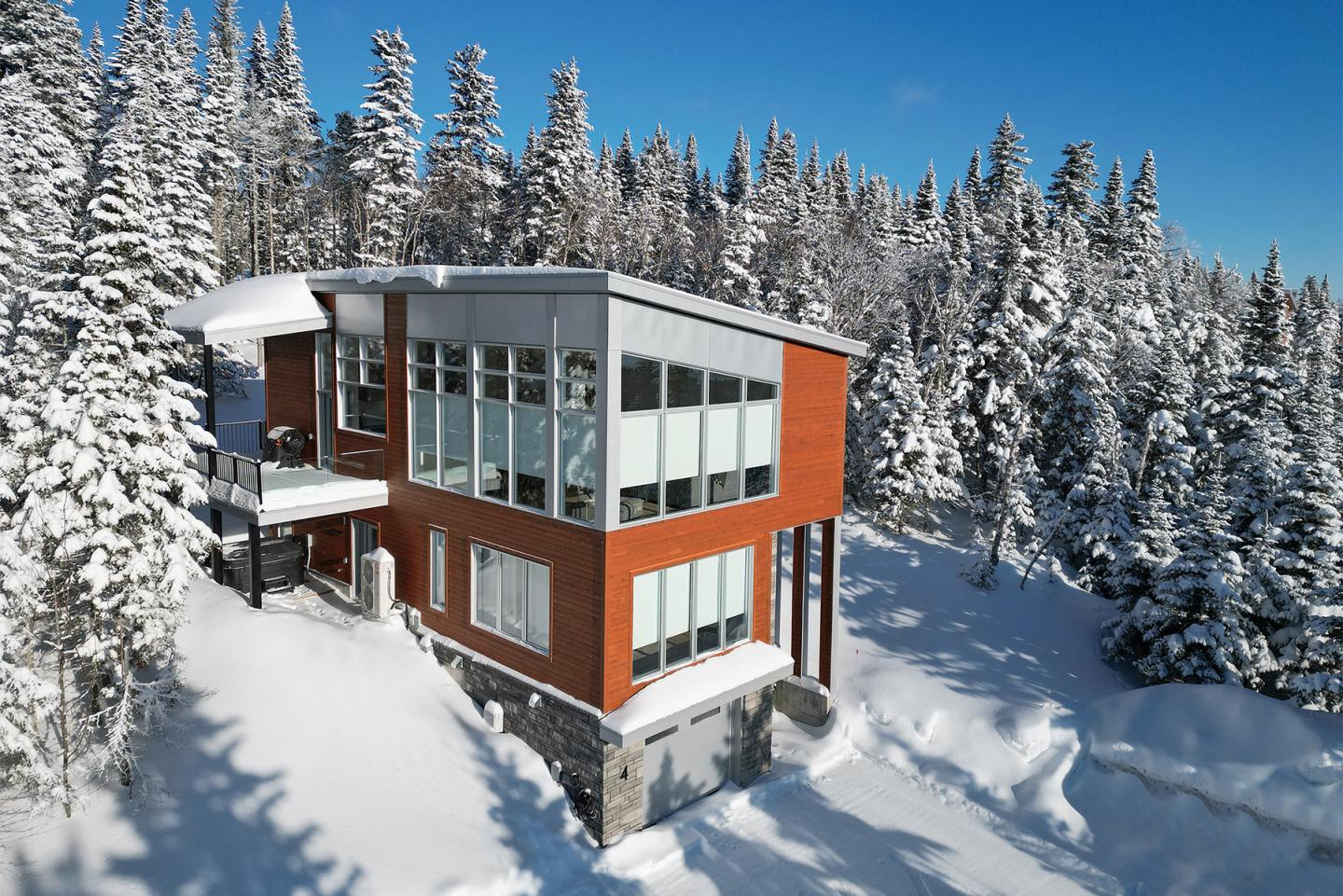 Maison moderne dans la neige entourée de sapins sous un ciel bleu.