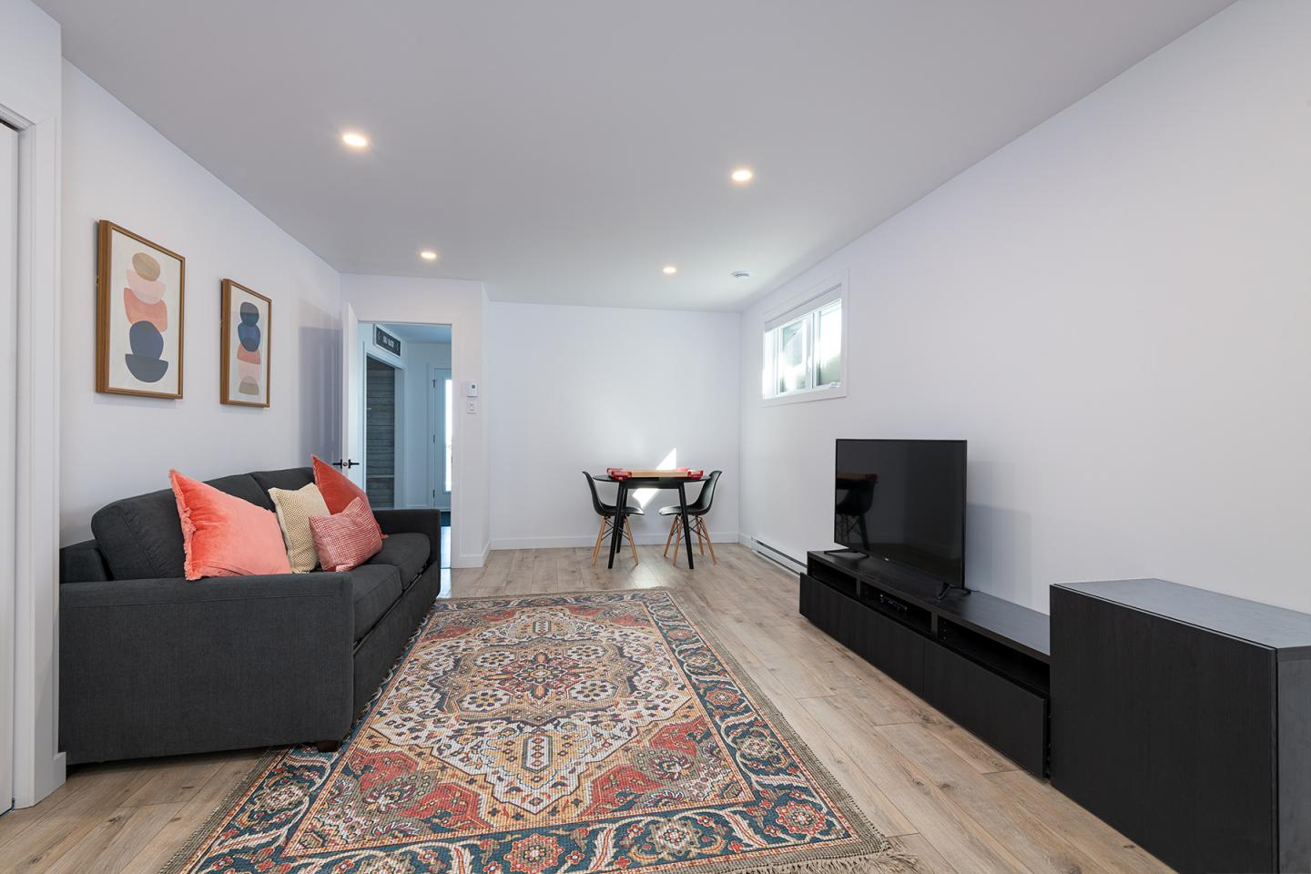 Salon moderne avec canapé noir, tapis coloré et télé, murs blancs, table et chaises au fond.