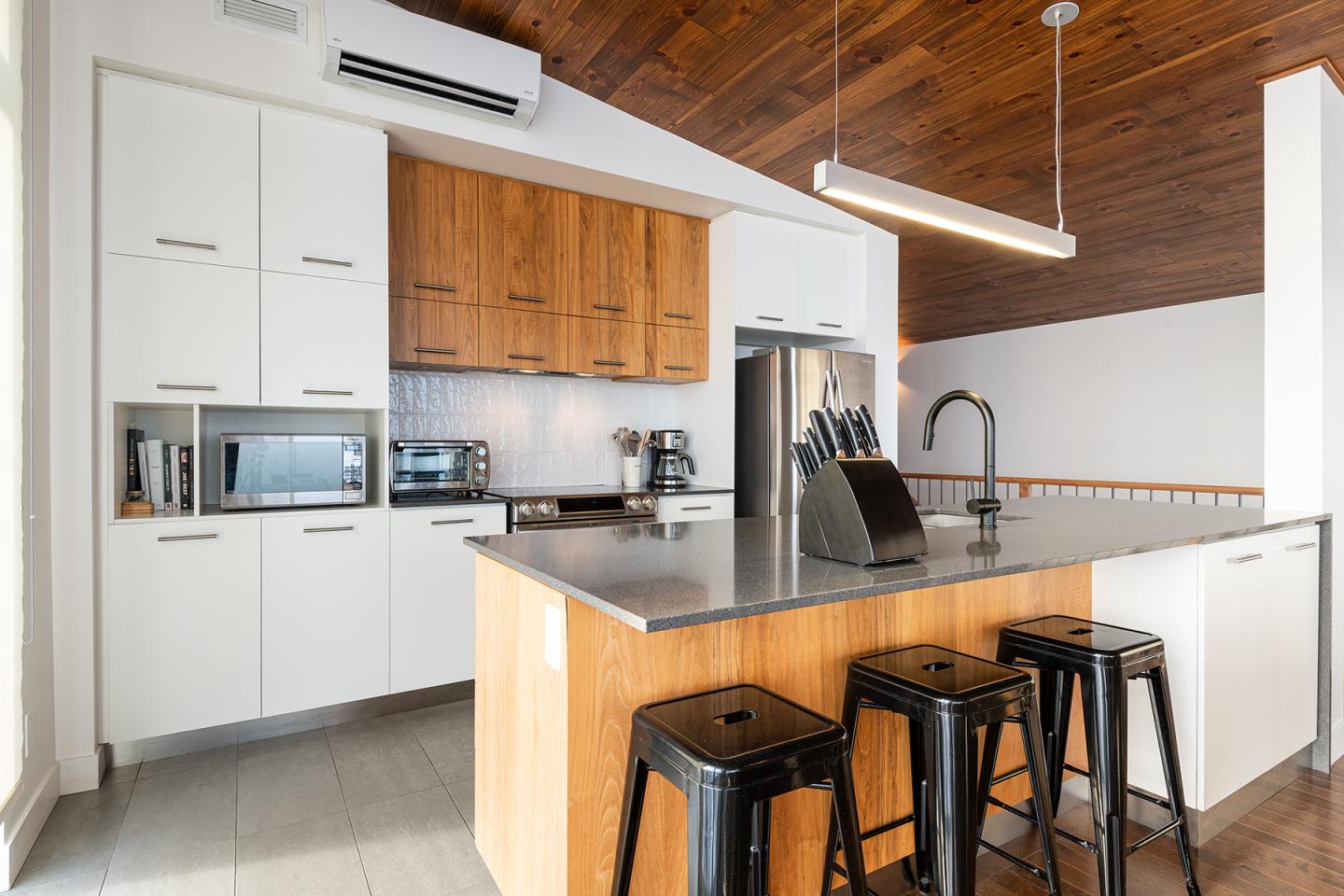 Cuisine moderne avec îlot central, tabourets noirs, et plafond en bois.