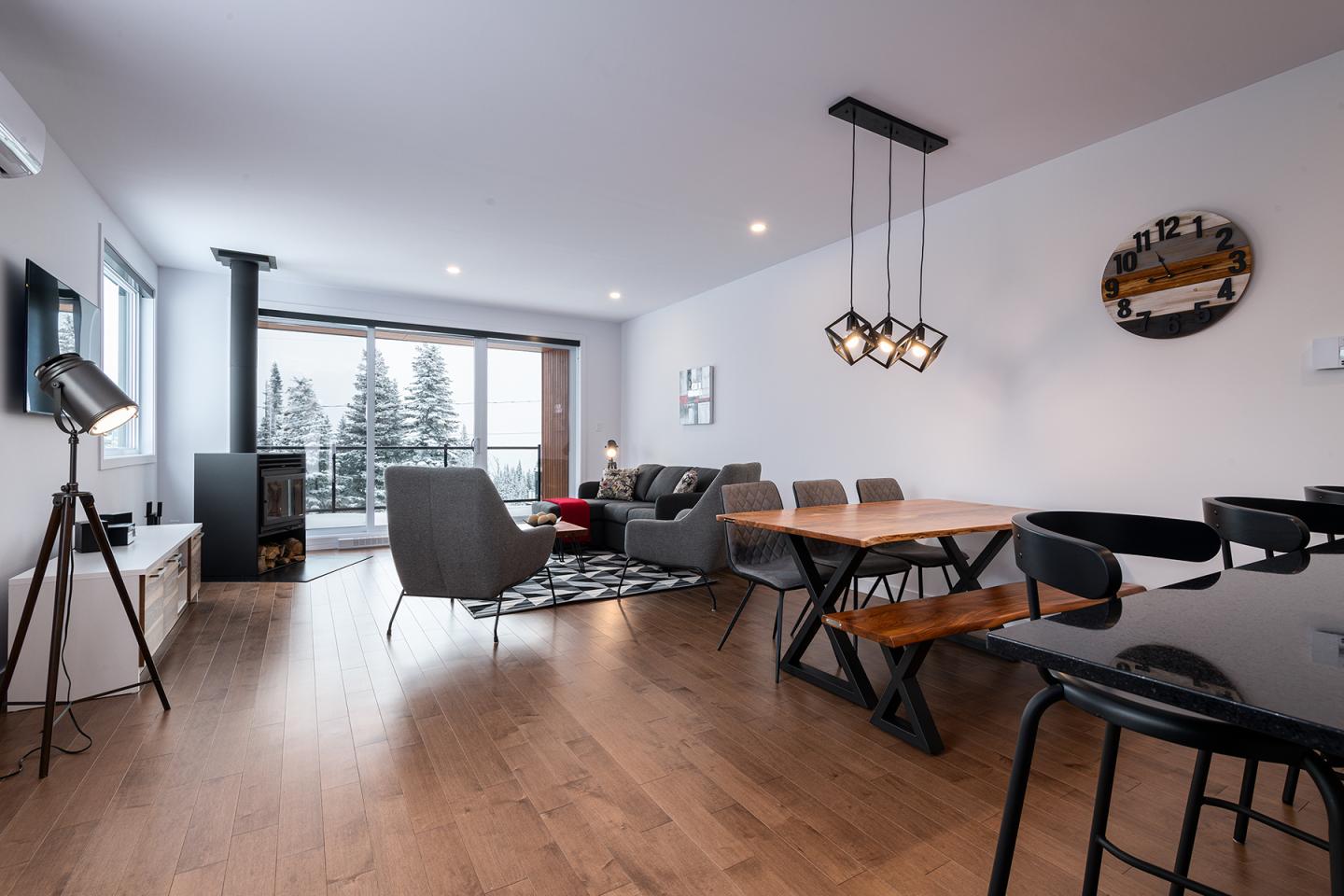 Salon moderne avec parquet, canapé, table en bois, et grandes fenêtres avec vue sur forêt enneigée.