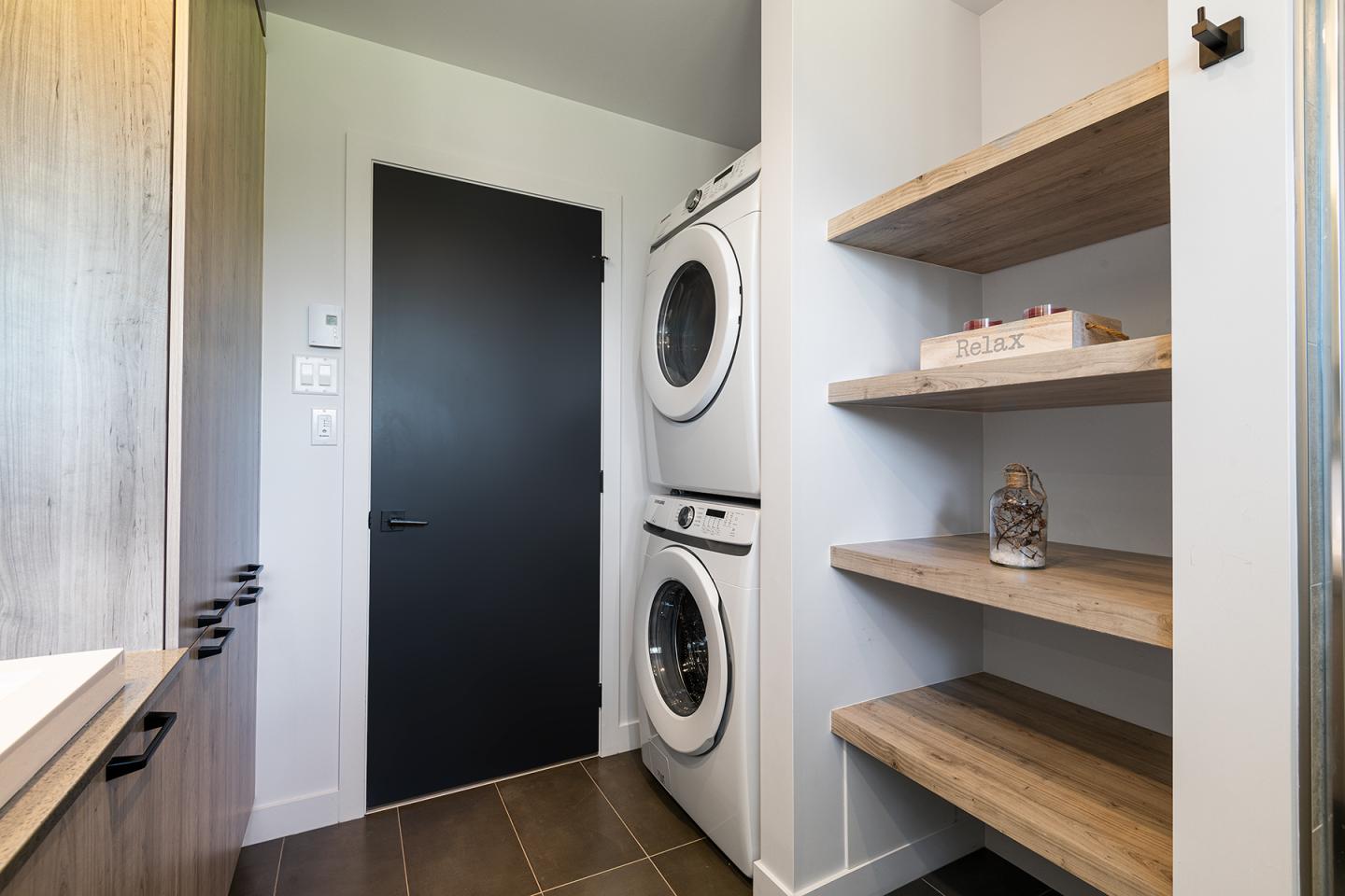 Salle de lavage avec lave-linge et sèche-linge empilés, étagères en bois à droite.