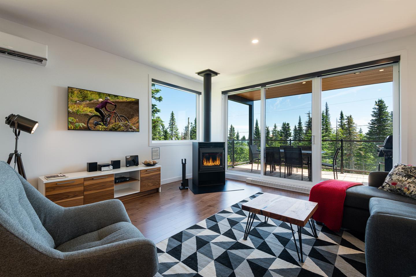 Salon moderne avec canapé gris, tapis géométrique, cheminée et vue sur la forêt.