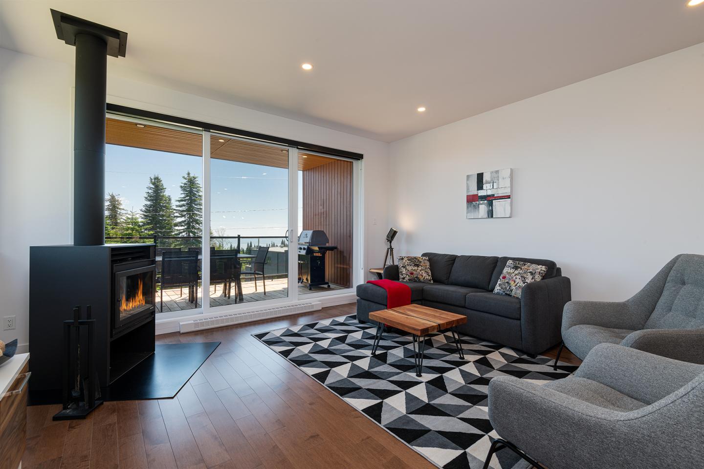 Salon moderne avec cheminée, canapé gris et grandes baies vitrées donnant sur des arbres.