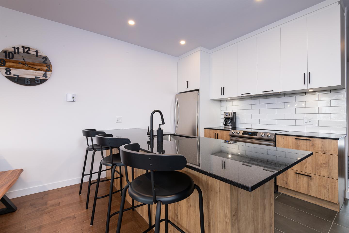 Cuisine moderne avec comptoir noir et tabourets, horloge murale, éclairage encastré.