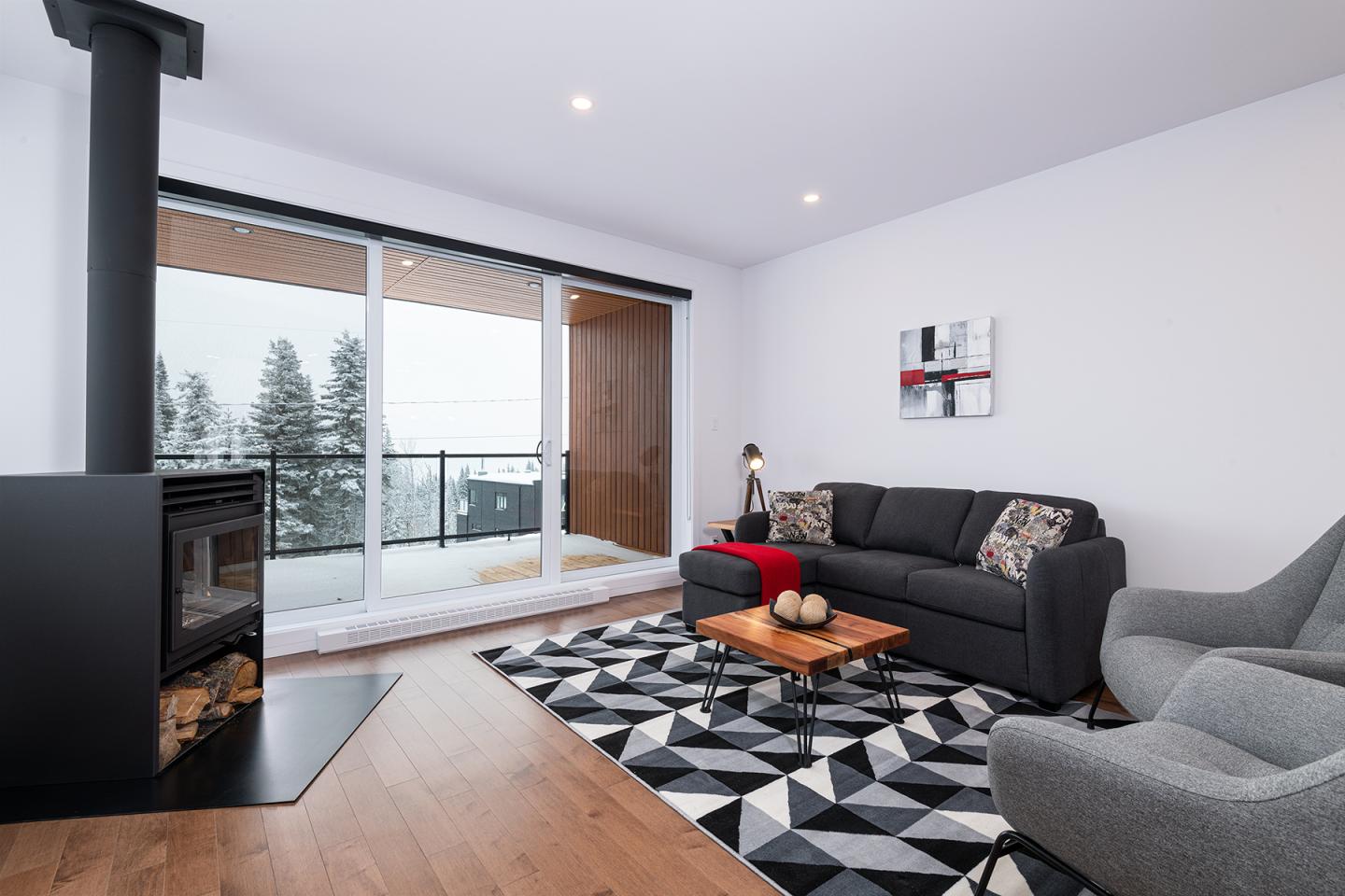 Salon moderne avec canapé gris, poêle à bois, grande fenêtre avec vue neige.