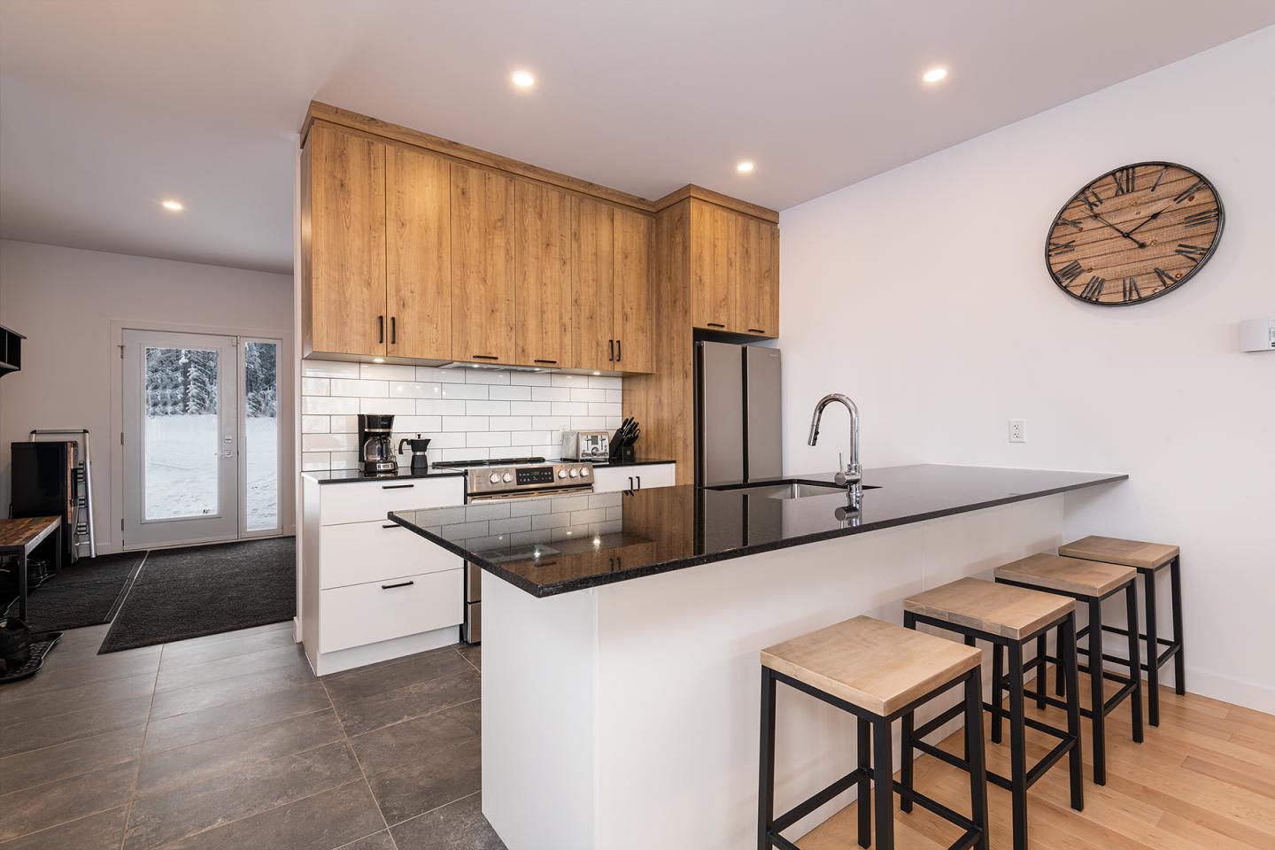 Cuisine moderne avec îlot noir, tabourets en bois, et armoires en bois clair. Horloge murale.