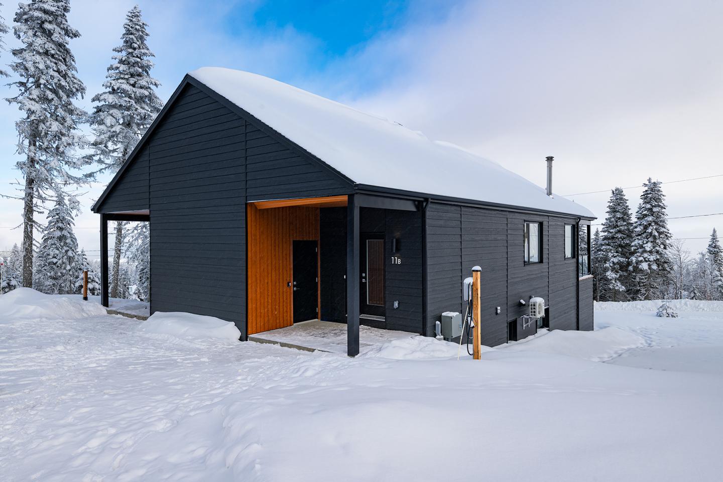 Chalet moderne noir dans un paysage enneigé, ciel bleu et arbres au fond.