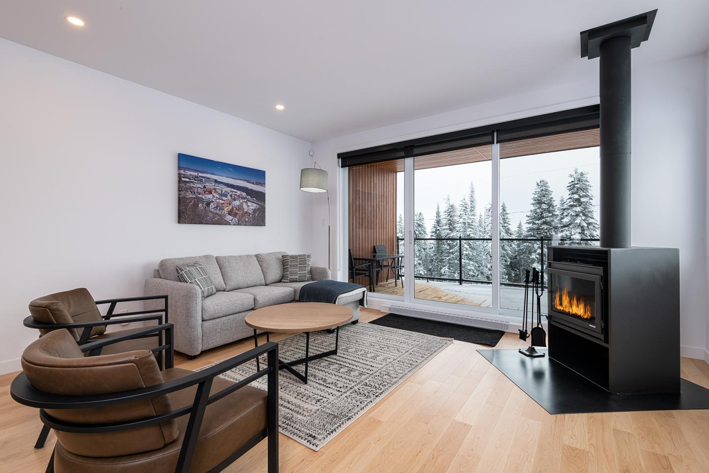 Salle de séjour moderne avec poêle à bois, canapé gris, et vue sur forêt enneigée.