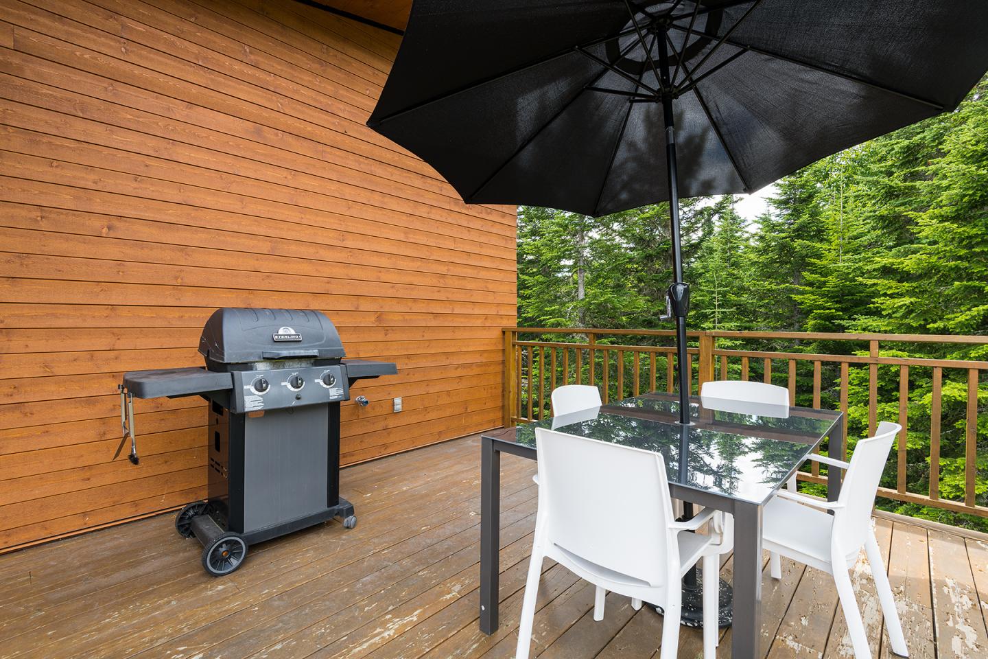 Terrasse en bois avec barbecue, table en verre, chaises blanches et grand parasol noir.