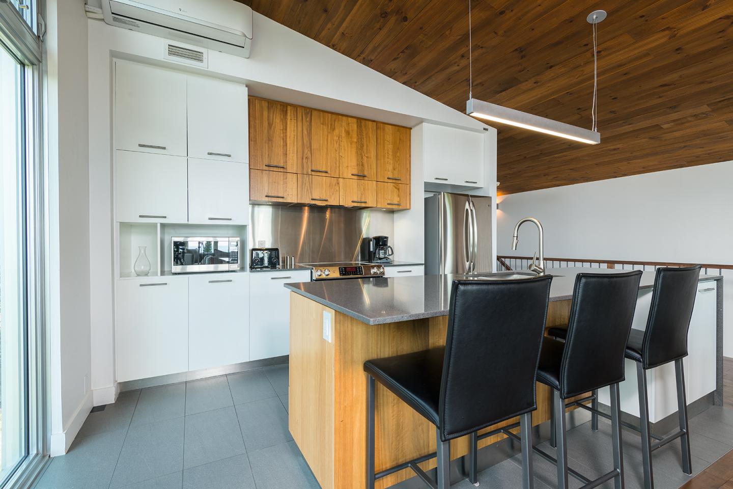 Cuisine moderne avec îlot central, chaises noires, placards en bois, sol et murs blancs.