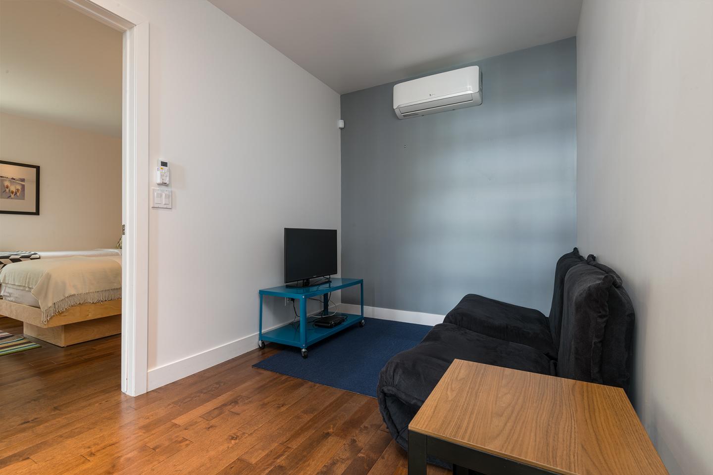 Salon minimaliste avec canapé noir, télé sur meuble bleu, plancher en bois. Ouverture sur chambre.