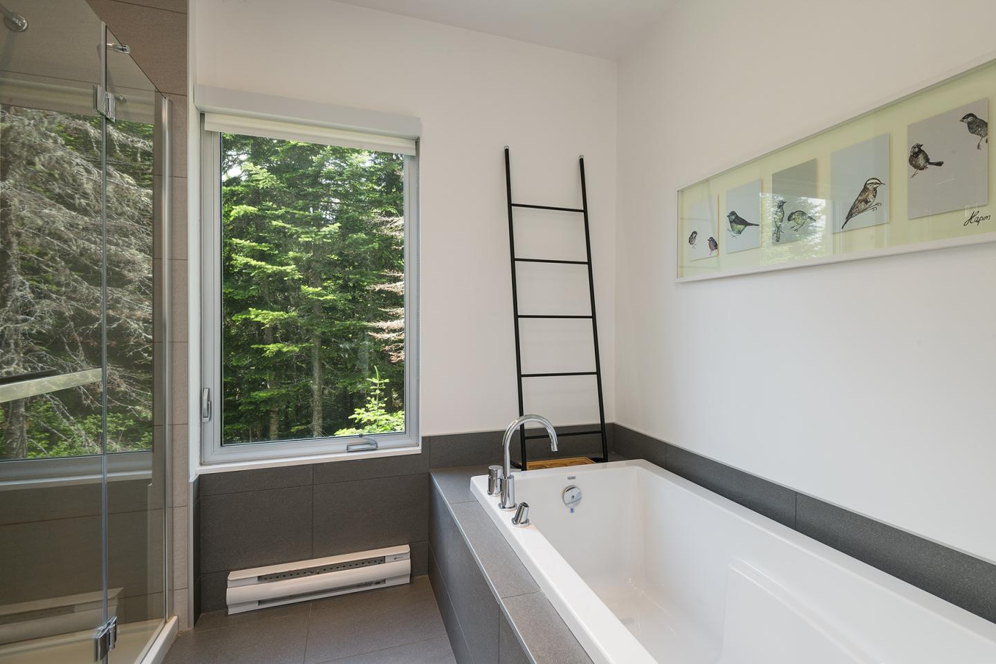 Salle de bain moderne avec baignoire, échelle noire, et fenêtre sur arbres.